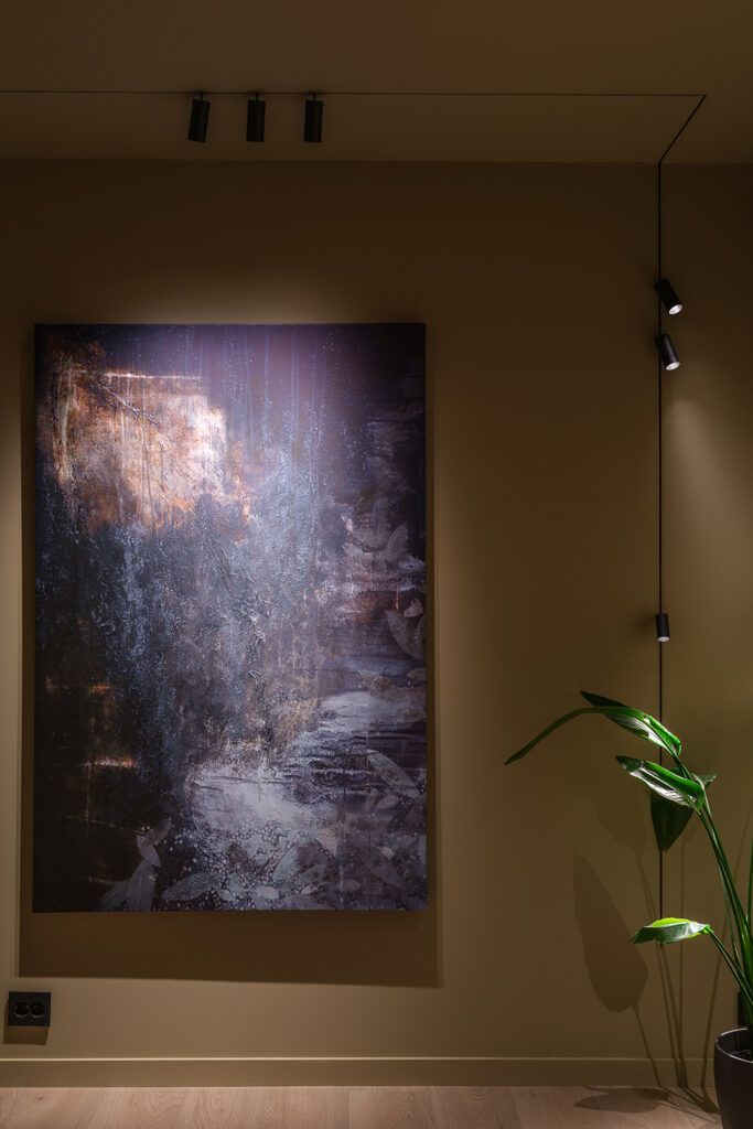 Et stort maleri på en vegg som blir belyst fra tre sorte spotter i taket.